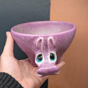 以下が含まれることがあります： ラベンダー色のセラミックボウルで、カバの顔のデザインが施されています。ボウルはまだらの質感で、青い目があります。カバの顔はボウルの前面にあり、耳と鼻が外側に伸びています。
