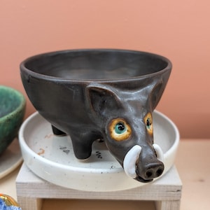 以下が含まれることがあります： ダークブラウンの釉薬が施された、イノシシの形をしたセラミックボウル。イノシシは白い牙と、緑、黄色、茶色のディテールを持つ目をしています。ボウルは、装飾的なデザインが施された白い皿の上に置かれています。