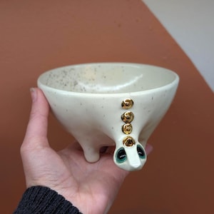 以下が含まれることがあります： 3つの小さな脚と装飾的な顔が付いた、斑点のあるオフホワイトのセラミックボウル。顔にはティール色の目と金色のアクセントがあります。ボウルは、茶色と白の背景に対して手で持たれています。