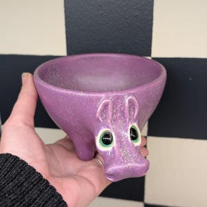 May include: A purple ceramic bowl shaped like a hippopotamus. The bowl has a speckled texture and features large, round eyes with green irises. The hippo has a small snout and two small ears. The bowl is held in a hand against a black and white checkered background.