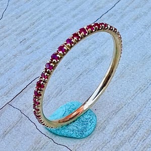 May include: A gold band ring with a row of small, red gemstones. The ring is displayed on a small, turquoise-colored stone against a light blue and gray surface. The ring's design is simple and elegant.
