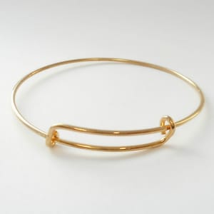 May include: A gold-toned bangle bracelet with a simple, open design. The bracelet is made of a single, thin wire that is bent into a circle and then joined at the ends with a small, decorative clasp.