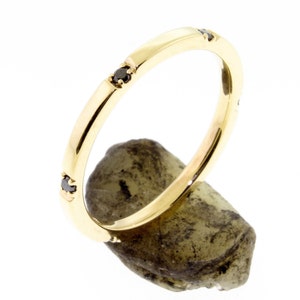 May include: A gold band ring with three small black gemstones set into the band. The ring is displayed on a textured, light green stone against a white background. The ring has a polished finish.