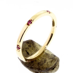 May include: A gold band ring with three small, round, red gemstones set into the band. The ring is displayed on a small, rough, gray-green stone against a white background. The ring has a polished finish.