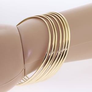 May include: A set of ten gold-toned bangle bracelets stacked on a mannequin arm. The bracelets are all the same width and have a smooth, polished finish.