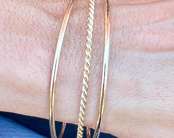 May include: Three gold bangles stacked on a wrist. The bangles are different widths and styles, including a twisted rope design.