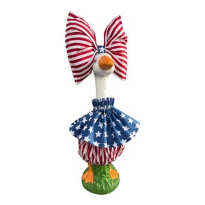 Könnte beinhalten: Dekorative Gänsefigur mit patriotischem Thema. Die Gans trägt eine große rot-weiß gestreifte Schleife, einen blauen Rock mit weißen Sternen und rot-weiß gestreifte Pumphosen. Die Gans steht auf einem grünen Sockel.