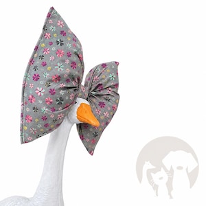 May include: A large, gray fabric bow featuring a repeating pattern of small pink, teal, and dark gray flowers. The bow is positioned behind the head of a white goose figurine with a bright orange beak, creating a decorative accessory display.