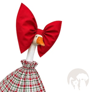 May include: A white goose figurine wearing a large, bright red fabric bow on its head and a ruffled dress featuring a red, white, and green plaid pattern. The goose has a distinct orange beak and a long, slender neck, with the fabric outfit covering its body.