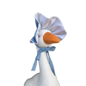 May include: A white goose figurine wearing a light blue and white fabric bonnet. The bonnet features a ruffled brim and matching light blue fabric ties knotted securely under the chin. The goose has a bright orange beak and a smooth white surface.