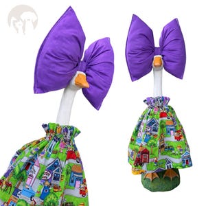 May include: A white goose figurine wearing a large purple fabric bow on its head and a green skirt featuring a colorful printed pattern of houses, trees, and small figures. The goose has an orange beak and stands on a green base.