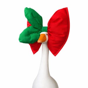 May include: A white goose figurine wearing a large fabric bow accessory. The bow features a bright red side and a green side with a leaf-like shape. The goose has a smooth white neck and a solid orange beak, with the bow positioned behind the head.
