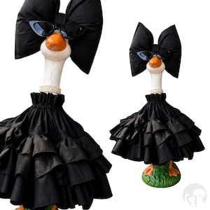 May include: A decorative goose figurine wearing a large black fabric bow on its head, black cat-eye sunglasses, a white pearl-style beaded necklace, and a black tiered ruffled skirt. The goose has a white neck and orange beak and feet.
