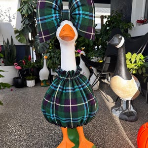 May include: A white goose statue wearing a green, blue, and purple plaid fabric outfit consisting of a ruffled skirt around its midsection and a large matching bow attached to its head. The goose has an orange beak and orange webbed feet.
