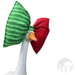 May include: A white goose figurine wearing a large fabric bow. The left side of the bow features a green striped watermelon rind pattern, while the right side is red with small black seed-like dots. The goose has a smooth white neck and orange beak.