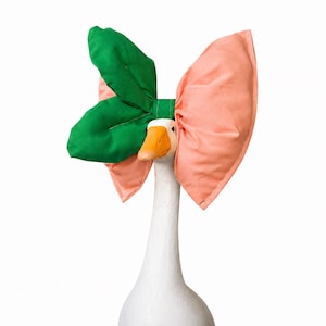 May include: A white goose figurine wearing a large, oversized fabric bow on its head. The bow features a vibrant green section on the left side and a soft peach-colored section on the right side, contrasting with the goose's bright orange beak.