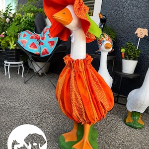May include: A white goose lawn ornament wearing a bright orange fabric outfit consisting of a gathered, polka-dot dress and a matching wide-brimmed sun hat. The goose has a painted orange beak and orange feet, standing on a green base.