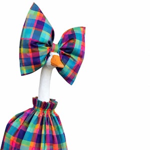 May include: A white goose figurine wearing a large, oversized bow and a ruffled skirt, both crafted from a vibrant, multi-colored plaid fabric featuring intersecting lines of blue, purple, green, orange, and pink against a white background.