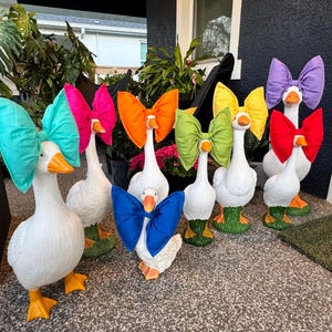 May include: A group of white goose ornaments, each with a large, brightly coloured bow. The bows are in shades of teal, pink, orange, green, yellow, red, and purple. The geese have orange beaks and feet, and some wear green wellington boots.