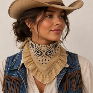 May include: A tan felt cowboy hat with a braided band, paired with a tan bandana featuring a black and white paisley print and a ruffled edge. The outfit includes a blue denim vest with brown fringe detailing and a light-colored button-down shirt.