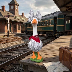 May include: A white goose figurine wearing a grey and white striped fabric outfit consisting of a large head bow and bloomers, accented with a red paisley bandana around its neck. The goose has an orange beak and orange webbed feet.