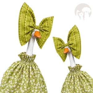 May include: Two white goose figurines with orange beaks, each wearing a green fabric outfit with a small white floral pattern and a gathered neckline. Each goose has a large, upright green bow with a white semi-circle pattern on its head.