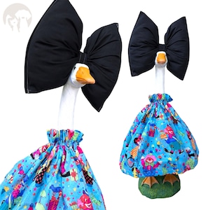 May include: A decorative goose statue wearing a large black fabric bow on its head and a blue dress featuring a colorful clown and gift pattern. The goose has a white neck, an orange beak, and stands on a green base with orange webbed feet.