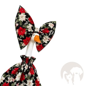 May include: A white goose figurine wearing a black fabric outfit featuring a pattern of red and white poinsettia flowers and green pine needles. The goose has a large matching fabric bow attached to its head and a gathered, ruffled neckline.