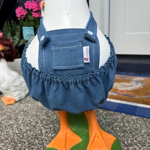 May include: A decorative goose statue wearing a blue denim apron with white buttons and a small pocket. The apron has a ruffled hem and a tag that reads "PetProperty". The goose has orange feet and stands on a green base.