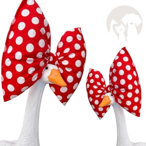 May include: Two white goose figurines, one larger than the other, each wearing a large, oversized fabric bow. The bows are bright red with a repeating pattern of white polka dots. Each goose has a distinct orange beak and a smooth white neck.