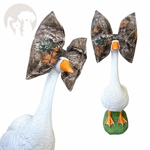 May include: A white goose figurine with an orange beak and feet, standing on a green base. The goose wears a large, oversized fabric bow featuring a brown, green, and tan camouflage pattern with printed leaf details on its head.
