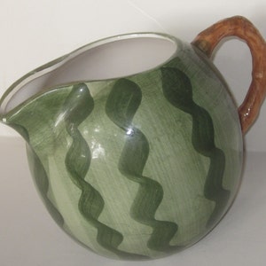 May include: Green ceramic pitcher with a brown wooden handle. The pitcher is shaped like a watermelon and has a green and white striped pattern.