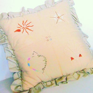 May include: A beige pillow with a ruffled edge and embroidered designs. The designs include a star, a flower, a heart, and a circle with dots.
