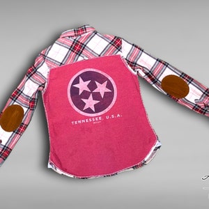 May include: A pink and white plaid flannel shirt with a red t-shirt underneath. The t-shirt has a purple circle with three white stars and the text "Tennessee, U.S.A." printed on the back. The shirt has brown leather elbow patches on both sleeves.