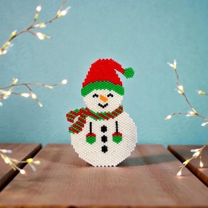 Puede incluir: Una figura de muñeco de nieve hecha de pequeñas cuentas blancas, rojas y verdes. El muñeco lleva un gorro rojo con un pompón verde y una bufanda a rayas verdes y rojas. Tiene una pequeña nariz naranja, ojos negros, boca negra y tres botones.