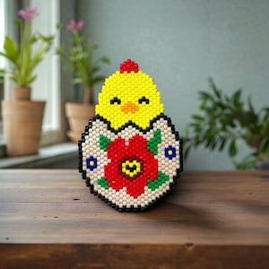 May include: A beaded Easter decoration featuring a yellow chick emerging from a decorated egg. The egg is adorned with a red flower and green leaves. The chick has a red comb. The decoration is on a wooden surface.