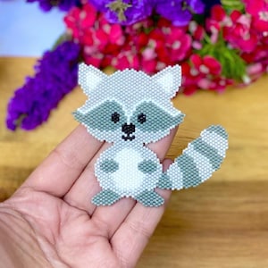 May include: A handmade beaded raccoon pin. The pin features a cute raccoon design with a striped tail and is made of small, gray, white, and black beads. The pin is a handmade item.