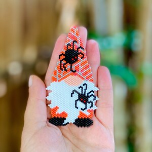 May include: A beaded gnome ornament with an orange and white hat and a black spider on its hat. The gnome is holding a black spider in its hand.