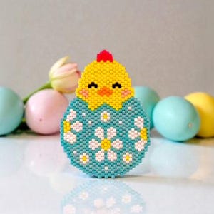 Peut inclure: Un poussin de Pâques en perles émergeant d'un œuf turquoise décoré de fleurs blanches. Le poussin est jaune avec une crête rouge et un jabot jaune. Des œufs de Pâques aux couleurs pastel sont en arrière-plan, sur une surface blanche.