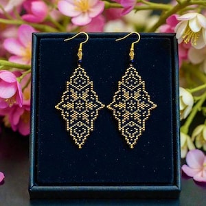 May include: A pair of gold and navy blue beaded earrings with a diamond-shaped pattern. The earrings are presented in a dark blue box, with pink and white flowers in the background. The earrings have gold-coloured hooks.