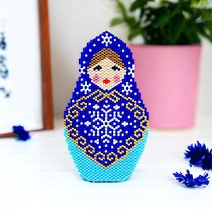 May include: A blue and white beaded Russian nesting doll, or matryoshka doll, with a snowflake pattern. The doll is wearing a blue and white hat and has a gold trim.