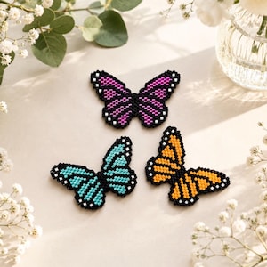 Pode incluir: Três decorações de borboletas de contas em cores vibrantes: roxo, turquesa e laranja. Cada borboleta tem um contorno preto e detalhes brancos. As borboletas estão dispostas sobre uma superfície clara, com detalhes florais.