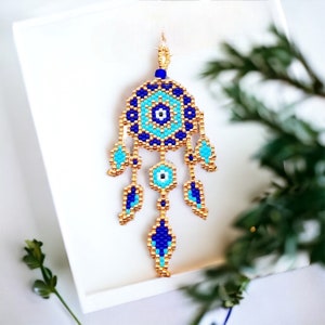 May include: A blue and gold beaded dreamcatcher with a white and blue evil eye charm. The dreamcatcher has three blue and gold beaded feathers.