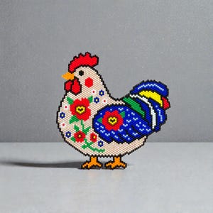 May include: A beaded rooster figurine with a beige body, red comb, and yellow beak. The rooster features red floral designs and a blue wing with yellow and green accents. The figurine is standing on a gray surface.