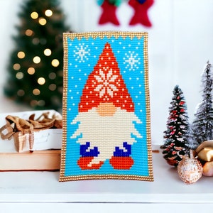May include: A beaded gnome ornament with a red hat and white snowflake design, against a blue background with white snowflakes. The gnome is wearing blue boots and has a white beard.