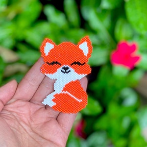 May include: A small, orange and white beaded fox with black eyes and a closed-mouth smile. The fox is sitting with its tail curled around its body.