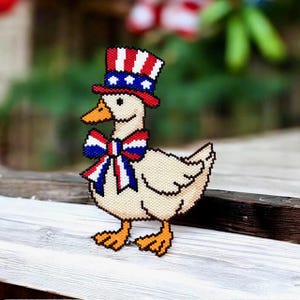 May include: A beaded goose figurine wearing a patriotic Uncle Sam hat and bow tie. The hat is red, white, and blue, with stars. The bow tie has a similar color scheme. The goose has orange feet and a beak, and is standing on a wooden surface.