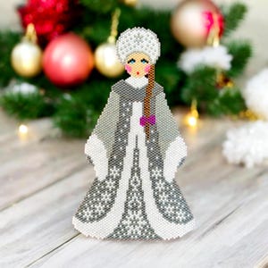May include: A beaded Christmas ornament depicting a woman wearing a white and gray dress with a white hat and a purple bow. The ornament is made of small beads and has a detailed design.