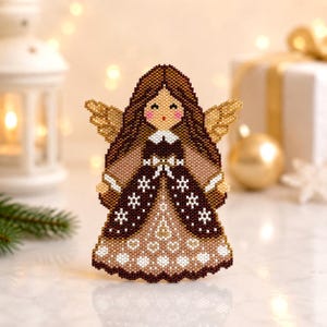 May include: A beaded angel ornament with brown hair, gold wings, and a brown and tan dress. The dress features snowflake and dot patterns. The angel has rosy cheeks and a white collar. The background includes a blurred gift box and Christmas lights.