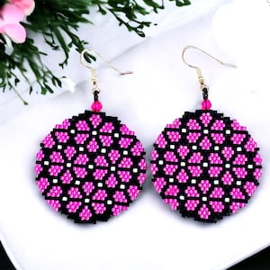 以下が含まれることがあります： 幾何学模様の黒とピンクのビーズで作られた丸いイヤリングのペア。イヤリングは丸く、シルバーのフックが付いています。